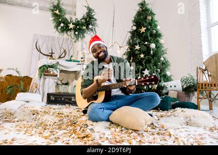 Bel ragazzo afroamericano in cappello di santa tiene giutaro e bengala luce, si siede sul pavimento di fronte alle decorazioni di Natale Foto Stock