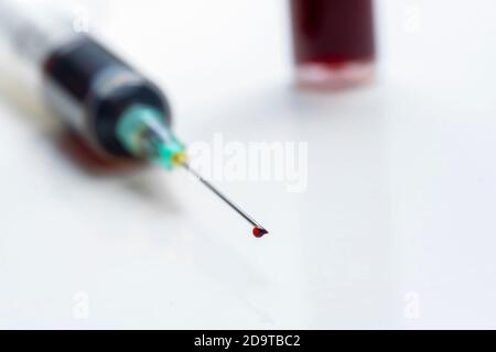 goccia di sangue sulla punta dell'ago di una siringa con una provetta per analisi del sangue non focalizzata sullo sfondo Foto Stock