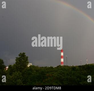 Dietro il verde denso sono un insediamento e un camino alto. Sopra il paesaggio si trova un cielo pesante e grigio con un arcobaleno Foto Stock