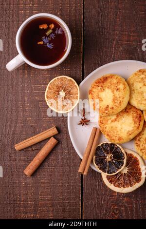 Frittelle di formaggio cottage con tè nero aromatico caldo, umore di prima colazione di Natale con anice, cannella e agrumi secchi su sfondo di legno, vista dall'alto Foto Stock