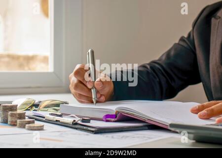 Una donna scrive in un notebook sul desktop con un mucchio di monete accanto alla finestra della casa e idee educative. Foto Stock