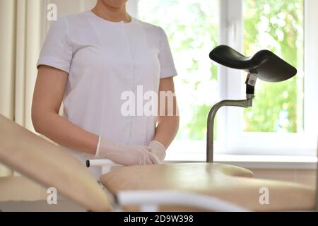 Ginecologa ragazza in tuta bianca e guanti in nitrile leggeri e protettivi si trova sulla sedia ginecologica. Foto Stock