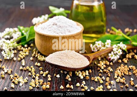 Farina di grano saraceno da cereali marroni in un cucchiaio, farina di grano saraceno da semole verdi in un recipiente, olio in un vaso di vetro, fiori e foglie sullo sfondo o Foto Stock