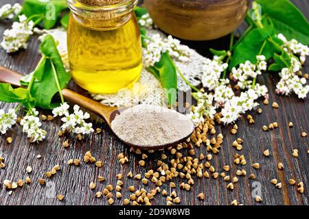 Farina di grano saraceno di cereali marroni in un cucchiaio, olio in un vaso di vetro su tela, fiori e foglie su sfondo di legno Foto Stock