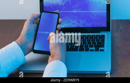 Le mani di un uomo navigano sullo schermo di un telefono cellulare di fronte a un computer portatile con la stessa immagine su una scrivania in legno. Luci blu Foto Stock