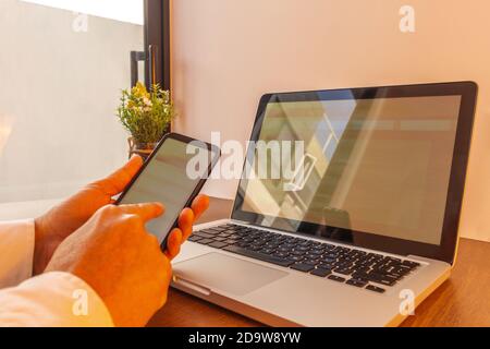 mani di un uomo che naviga attraverso il suo cellulare con un computer portatile coordinato con lo stesso sito web. Immagine in toni caldi su una scrivania in legno in un Foto Stock