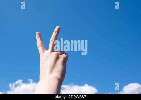 Mano con due dita in alto nel simbolo della pace o della vittoria. Anche un segno per la lettera V in lingua dei segni. Isolato su sfondo blu cielo con nuvole, copia spazio. Foto Stock