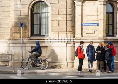 RIGA, LETTONIA. 19 ottobre 2020. Un uomo che indossa una maschera protettiva per il viso corre in bicicletta, mentre i giovani si trovano vicino al crosswalk. Foto Stock