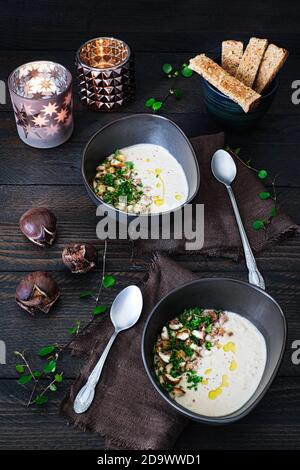 Due identiche zuppe di castagne cucinate di fresco in ciotole di ceramica marrone, decorate con pezzi di castagno e prezzemolo su un tavolo di legno marrone con sale e p Foto Stock