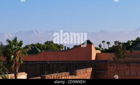 Montagne innevate dell'Atlante sopra la nebbia vista dallo storico Palazzo El Badi con costruzioni in legno e palme a Marrakech. Foto Stock