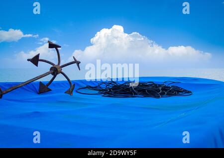 Anchor arrugginito e corda nera su una barca da pesca coperta in telo di telone blu su una spiaggia con blu nuvoloso cielo sullo sfondo Foto Stock