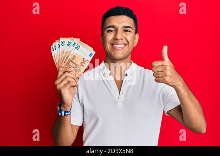 Giovane arabo che tiene 50 banconote in euro sorridendo felice e positivo, il pollice su facendo eccellente e segno di approvazione Foto Stock