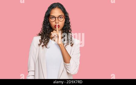 Giovane afroamericana che indossa abiti da lavoro chiedendo di essere tranquillo con il dito sulle labbra. Silenzio e concetto segreto. Foto Stock