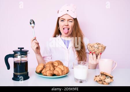 Giovane bella donna rossa seduta sul tavolo con la colazione che attacca la lingua fuori felice con l'espressione divertente. Foto Stock
