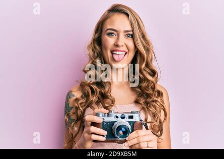 Giovane ragazza bionda che tiene vintage macchina fotografica attaccando lingua fuori felice con l'espressione divertente. Foto Stock