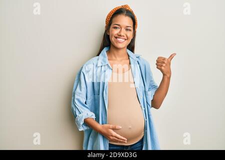 Bella donna ispanica in attesa di un bambino, toccando la pancia incinta puntando alla schiena dietro con mano e pollici su, sorridente sicuro Foto Stock