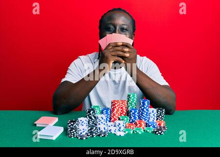 Bel giovane uomo nero che gioca a poker coprendo faccia con carte sorridenti con un sorriso felice e fresco sul viso. Mostrando denti. Foto Stock