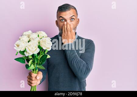 Bell'uomo di mezza età che tiene bouquet di fiori bianchi che coprono la bocca con la mano, scioccato e impaurito per errore. Sorpresa espressione Foto Stock