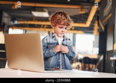 Bambino intelligente in abiti casual con computer portatile sul tavolo hanno divertimento con la lente d'ingrandimento Foto Stock