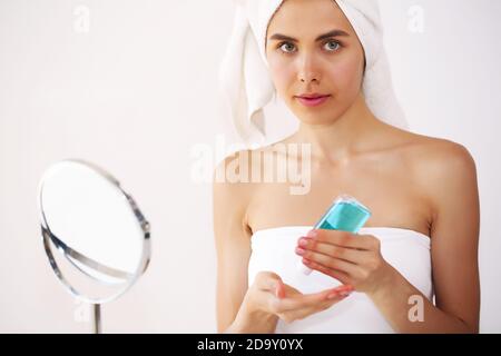 La giovane bruna si prende cura della sua pelle, in piedi davanti ad uno specchio, godendo di trattamenti di bellezza per se stessa, sorridendo teneramente Foto Stock