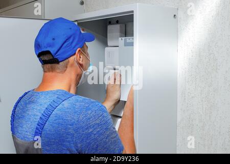 Un elettricista in tute e un cappello da baseball esamina un cabinet con sensori e attrezzature. Foto Stock