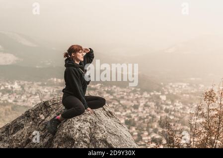 Bella ragazza dai capelli rossi è seduta sulle pietre sopra il precipizio contro lo sfondo di montagne e nebbia. Foto Stock