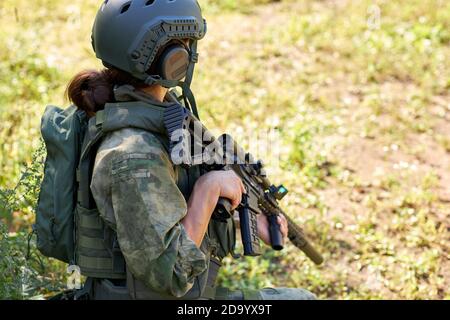 vista laterale su donna irriconoscibile in abbigliamento militare seduto sull'erba in attesa di nemico a lato, andando a sparare Foto Stock