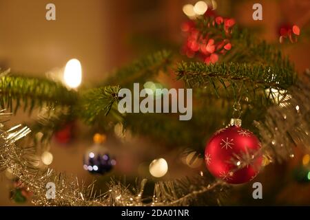 Primo piano di un ramo di albero di Natale con candele brucianti, corona, tinsel colorati baubles al coperto in serata. Colore classico di Natale verde, rosso Foto Stock