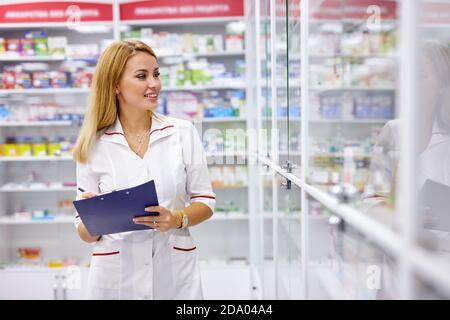 donna bionda dricgista in uniforme controllando le scorte di assortimento in farmacia, leggere le indicazioni Foto Stock