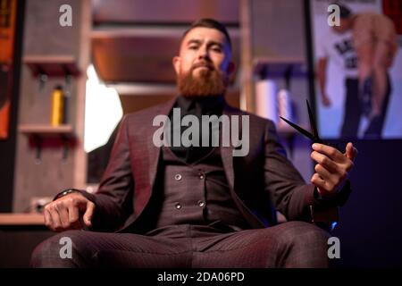 lo stylist professionale dei capelli è pronto per tagliare i vostri capelli o barba, maschio in abito sul posto di lavoro, in posa in stanza buia Foto Stock