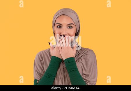 Madonna musulmana in Hijab coprendo la bocca con le mani, sfondo giallo Foto Stock