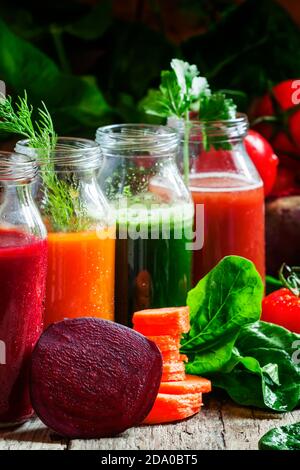 Quattro tipi di succhi di verdure: Rosso, borgogna, arancio, verde, in piccole bottiglie di vetro, verdure fresche ed erbe, sfondo di legno d'annata, selettivo Foto Stock
