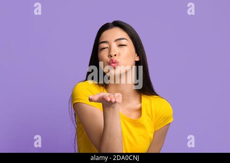 Carino giovane donna asiatica soffia aria bacio a macchina fotografica sopra Sfondo viola Foto Stock
