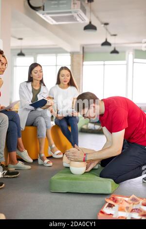 Classe CPR con giovani istruttori caucasici che parlano e dimostrano aiuto al primo soccorso, manichino cpr sul pavimento Foto Stock