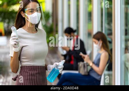 Ritratto attraente cameriera asiatica indossare maschera viso e scudo tenere termometro e igienizzatore gel di alcol con sfondo di cliente di distanza sociale Foto Stock