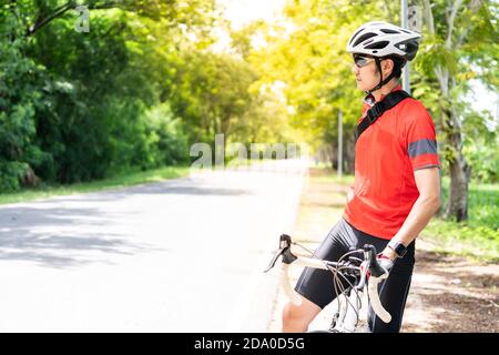 Ritratto del ciclista maschile asiatico in camicia sportiva con la sua bicicletta in strada di campagna con albero verde sullo sfondo. Weekend sportivo all'aperto atleta A. Foto Stock