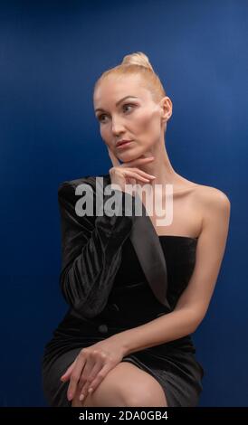 Fortunato giovane seduto su una sedia, meraviglioso in un abito da smoking con labbra delicate sfondo blu con luce studio. FLAUNTS Foto Stock