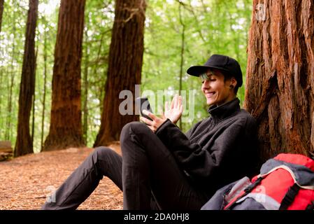 Vista laterale della vivace turista femminile che pende sull'albero dentro woods e videochiamare tramite smartphone mentre si agitano le mani E riposando a Monte Cabezon Natu Foto Stock