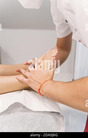 Vista laterale del terapeuta maschile che fa massaggio dei piedi per paziente irriconoscibile giacente sul tavolo in sala medica durante la riabilitazione sessione Foto Stock