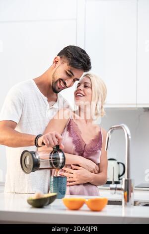Coppia amorevole in pigiama abbracciando e versando caffè fresco dentro durante la colazione, riunitisi mentre si sta al tavolo in cucina di mattina Foto Stock