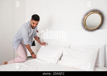 Uomo calmo indossando abiti domestici rendendo morbido letto in accogliente camera da letto dopo il risveglio di mattina Foto Stock