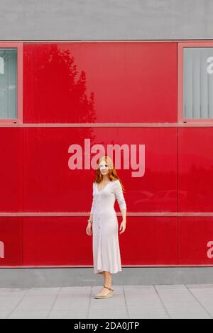 Corpo pieno di giovane femmina rosso in elegante abito beige in piedi sul marciapiede contro il luminoso muro rosso della moderna città edificio Foto Stock