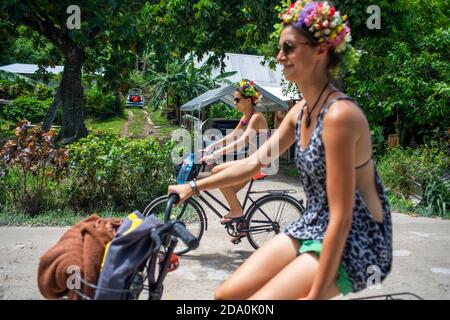 Due donne che cavalcano la sua bicicletta lungo una strada con palme a Bora Bora, Isole della Società, Polinesia francese, Sud Pacifico. Sud Pacifico Foto Stock