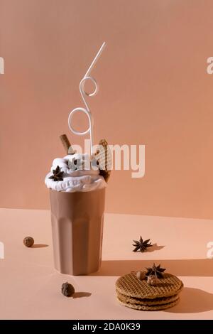 Bicchiere con cocktail al cioccolato con panna montata guarnita con stella anice e bastone di cannella messi a tavola con nocciole e. biscotti su bro pastello Foto Stock
