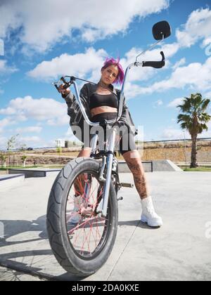 Da sotto determinata femmina tatuata in outfit elegante seduta sopra BMX in bicicletta nello skate Park e guardando la macchina fotografica Foto Stock