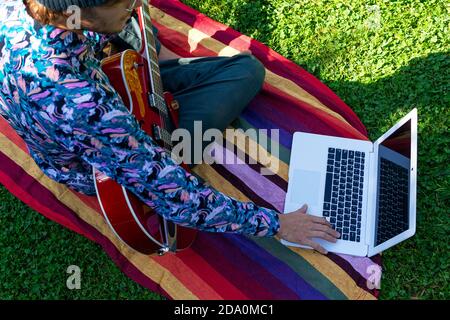 Dall'alto troncato irriconoscibile musicista maschile seduto su coperta con computer portatile e scelta di tutorial mentre si prepara per giocare moderno elettrico guita Foto Stock