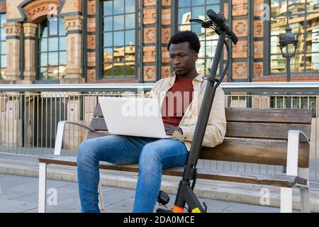 Freelance africano-americano concentrato seduto su panca vicino elettrico scooter e lavorando al progetto mentre si usa il netbook in città Foto Stock