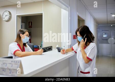 Infermiera femminile concentrata che indossa tuta medica bianca e maschera facciale prendere appunti nella cartella clinica mentre ci si avvicina al moderno accoglienza ospedaliera Foto Stock