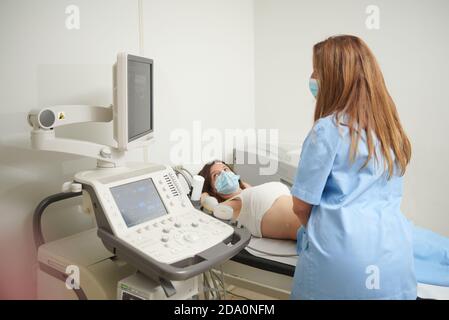 Anonimo medico femminile in uniforme esame paziente in maschera mentre monitoraggio del monitor dello scanner a ultrasuoni in clinica Foto Stock