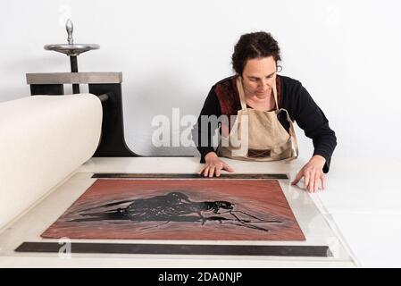 Focalizzato medio invecchiato femmina master mettendo foglio di linoleum con ornamento inchiostrato sulla pressa da stampa mentre si lavora in tecnica di linocut in studio Foto Stock